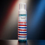A bottle of Barber Glaze foaming lotion stands centered on a dark surface with dramatic red and blue lighting in the background. The product is a clear plastic pump bottle with a white cap and a white label featuring red and blue horizontal stripes.